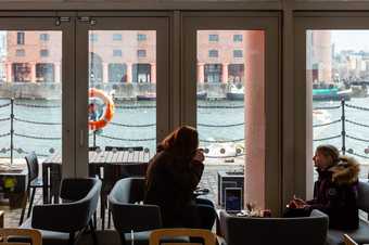 2 people are sat on sofas having drinks on a coffee table. Behind there are large windows which look onto the water in the Albert Dock. 